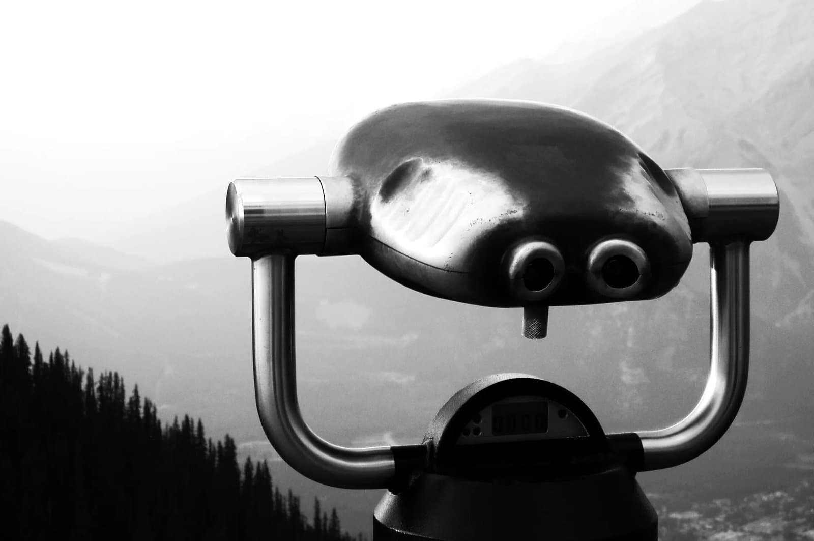 close‑up of a metal viewing binocular on a lookout point, with mountains blurred in the background.