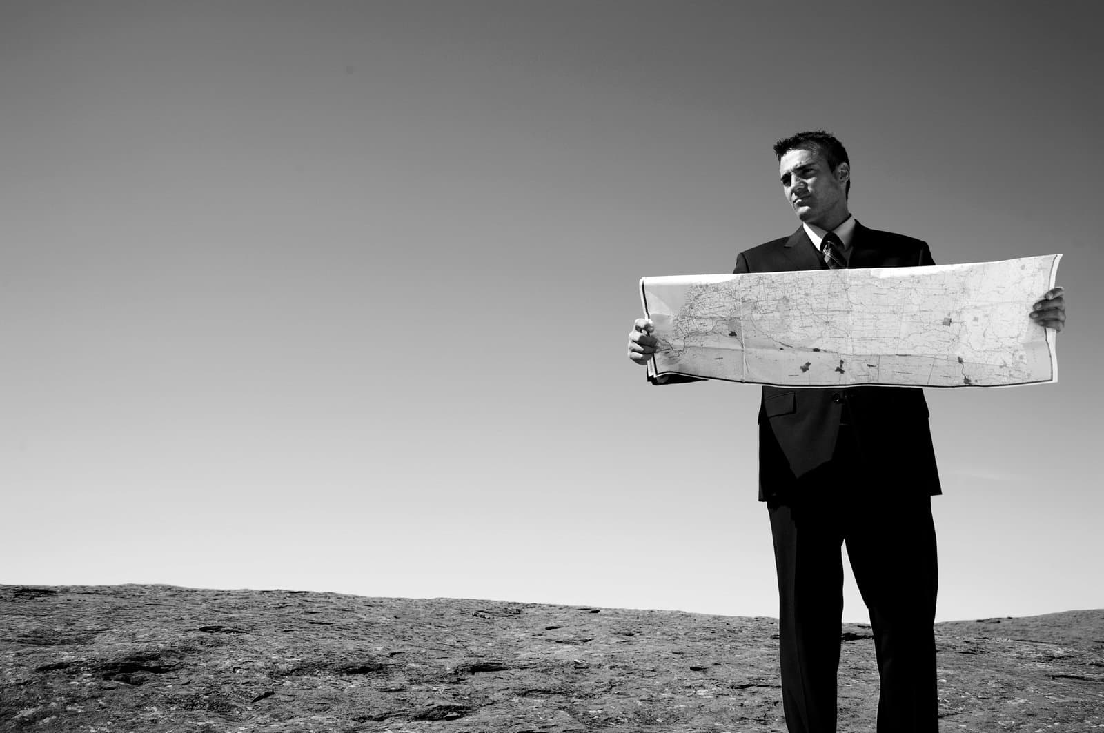 A businessman, lost in the desert, consults his map. He is looking off into the empty copy space to image left.