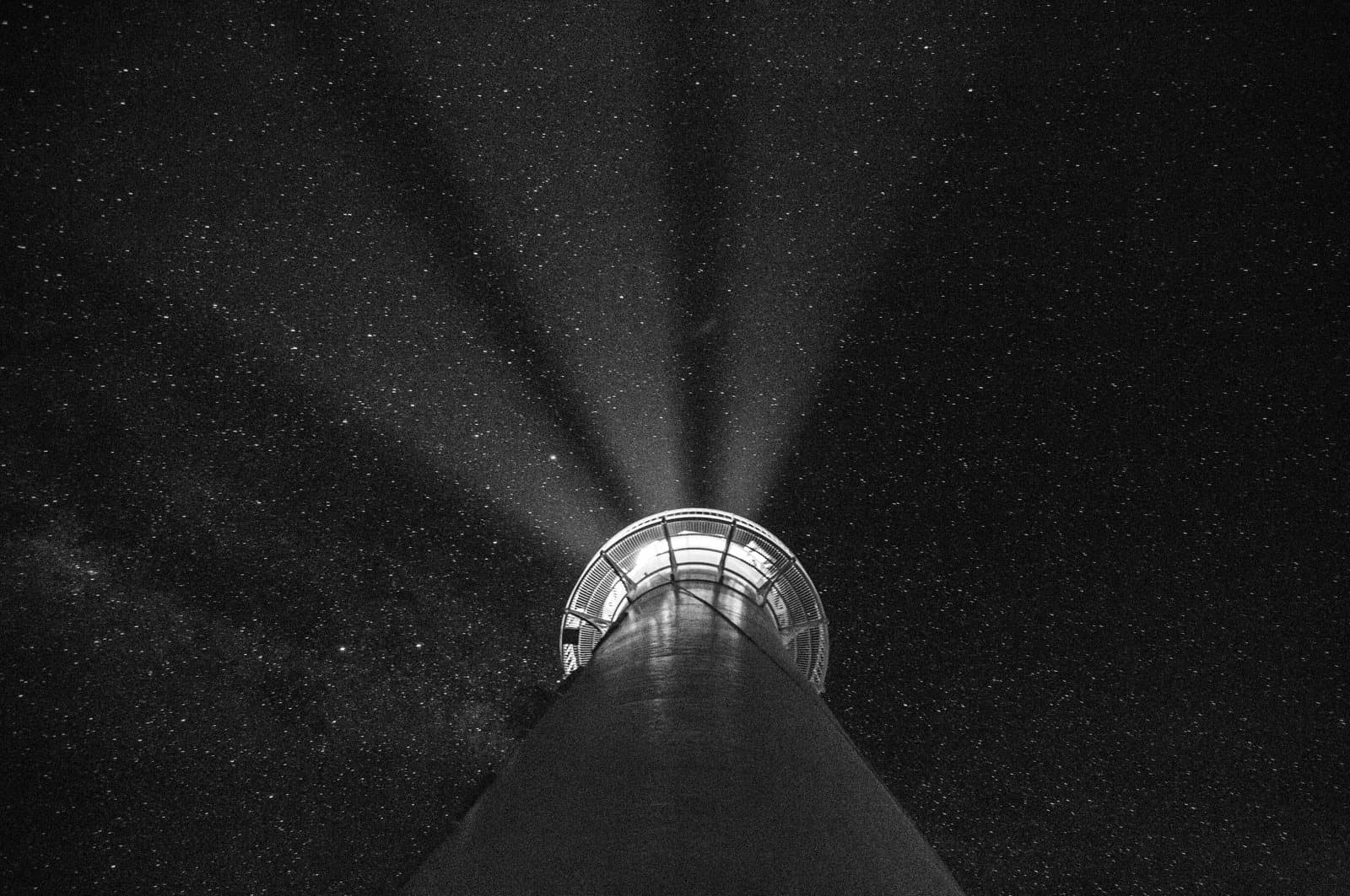 Lighthouse at night with beams of light radiating upward against a star‑filled sky.
